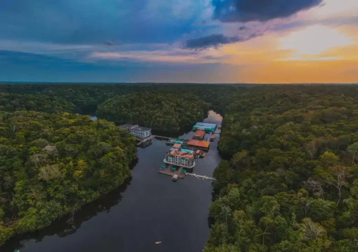 Vista aérea de um hotel flutuante em um rio cercado por uma floresta tropical exuberante ao pôr do sol, destacando-se por sua localização na natureza.