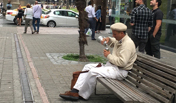 Homem vestido com roupa branca e chapéu sentado em banco de praça, tomando chimarrão, em uma rua movimentada com pessoas e carros ao fundo.