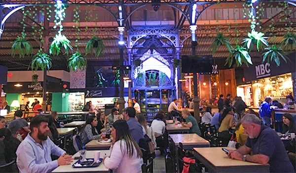 Ambiente movimentado de restaurante ou praça de alimentação, com várias pessoas sentadas em mesas, iluminação moderna, plantas suspensas e detalhes arquitetônicos sofisticados.