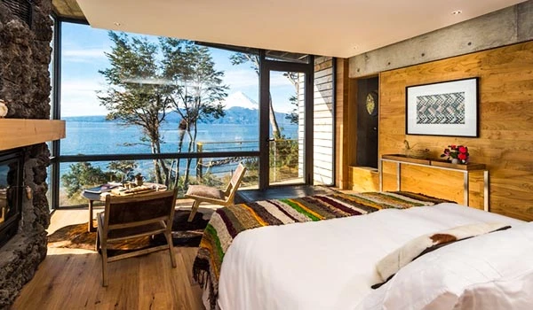 Vista de um quarto de hotel com decoração rústica e vista panorâmica para o lago e árvores através de grandes janelas, com uma cama de casal, mesa de jantar e cadeiras.