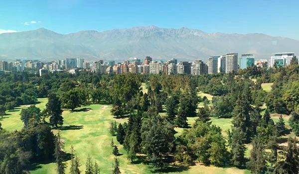 Vista panorâmica de um parque com árvores e espaço verde, com uma cidade ao fundo e uma cadeia de montanhas ao horizonte, sob céu azul claro. Lua de Mel no Chile.