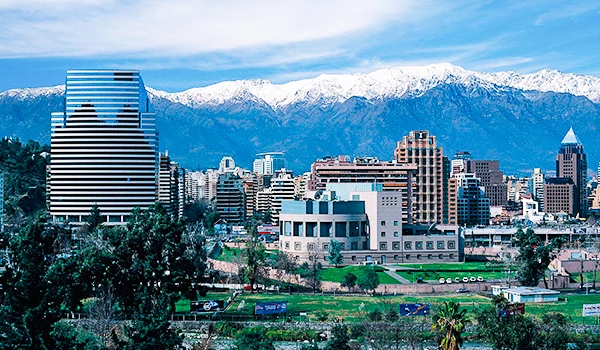 Vista panorâmica de um skyline urbano com edifícios modernos, ao fundo a cordilheira dos Andes coberta de neve, e um parque verde na cidade.