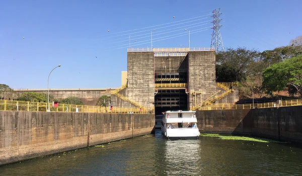 Represa de usina hidrelétrica com passagem de barco pelo vertedouro, mostrando estrutura de concreto e área de controle de água, cenário de produção de energia limpa.