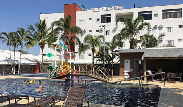 Piscina de hotel com pessoas se divertindo e área de lazer com cadeiras na frente de prédio de apartamentos com muitas palmeiras ao redor em um dia ensolarado.