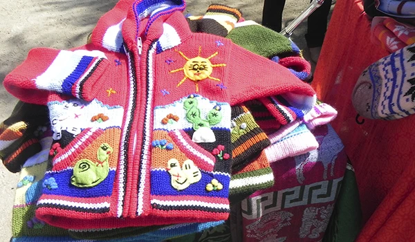 Casaco infantil de lã colorido com detalhes de sol, nuvem, estrela, caracol e outros desenhos, exposto em feira de roupas infantis ao ar livre.