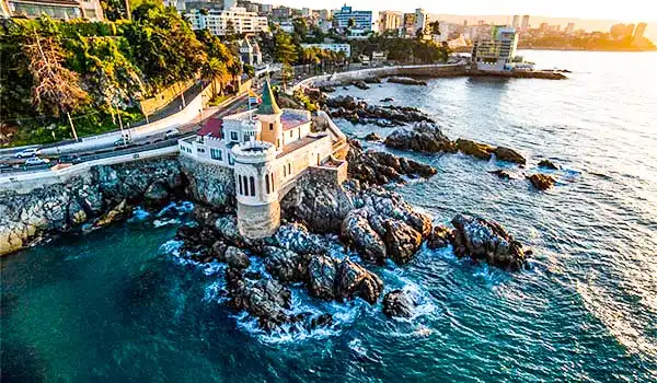 Vista panorâmica do litoral com uma torre histórica localizada na rocha, cercada pelo mar e urbanização ao fundo, ao pôr do sol, destacando o cenário de uma costa famosa. Viña del Mar o que fazer.