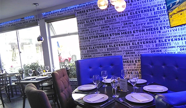 Interior de restaurante decorado com paredes azuis, mesas bem postas com louças e taças de vinho, iluminação moderna e decoração acolhedora.