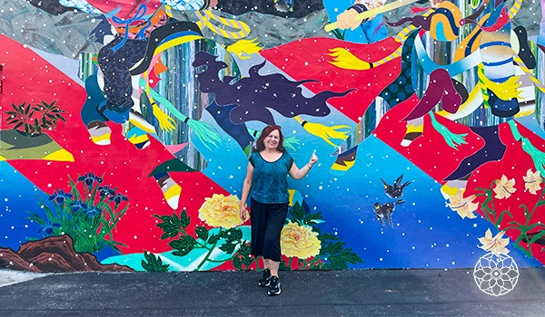 Deusa Rodrigues posando diante de um mural colorido com elementos artísticos, flores e animais, representando a beleza da arte urbana adaptada ao cenário contemporâneo.
