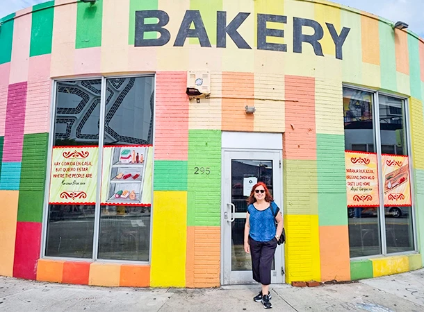 Deusa Rodriuges de cabelo castanho e óculos escuros na frente de uma padaria colorida com paredes pintadas em várias cores, destacando a palavra 'BAKERY'.
