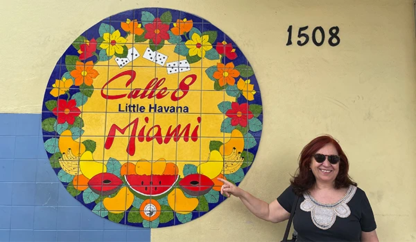 Deusa Rodrigues apontando para o mural colorido de Calle 8 em Miami, destacando a cultura vibrante cubana e a famosa rua. Material artístico e humor na foto.