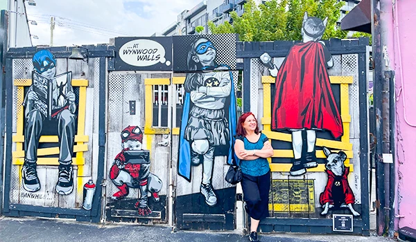 Mural de arte de rua com personagens de cachorro super-heróis em um grafite colorido, Deusa Rodrigues posando na frente na parede de uma rua urbana. O que fazer em Wynwood, Miami.