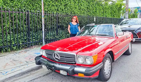 Deusa Rodrigues ao lado de um carro clássico Mercedes-Benz vermelho, estacionado na rua perto de uma cerca de ferro, com fundo de árvores e céu claro. O que fazer em Wynwood, Miami.