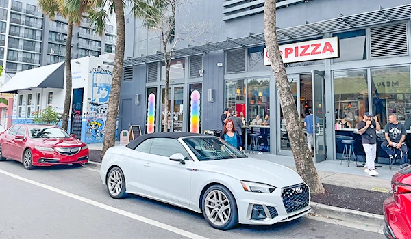 Imagem de uma pizzaria moderna localizada em uma rua movimentada, com carros estacionados na frente e pessoas ao redor. A fachada da pizzaria tem um letreiro visível e um ambiente urbano com árvores ao lado.