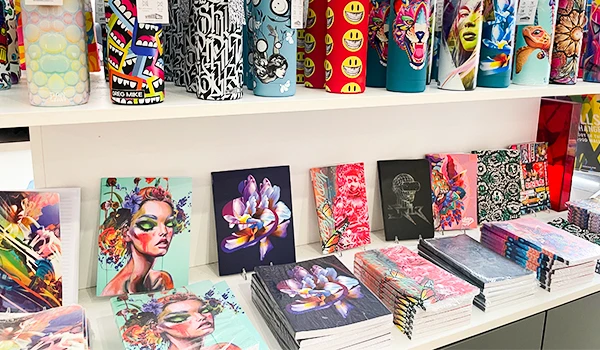 Diversos produtos de papelaria com estampas coloridas e artísticas, incluindo cadernos, livros e copos com designs vibrantes e criativos. Ideal para estudantes e amantes de arte.