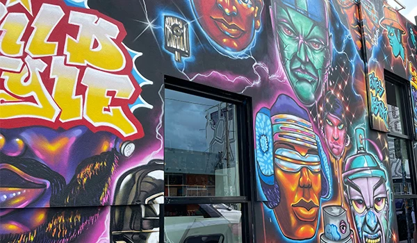 Entrada do Museum of Graffiti decorada com arte de rua vibrante, incluindo uma mulher com cabelo black, rosto rosa, e máscaras coloridas ao redor, destacando a cultura urbana.