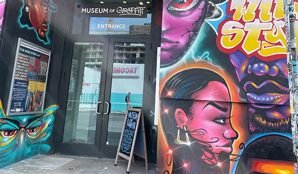 Entrada do Museum of Graffiti decorada com arte de rua vibrante, incluindo uma mulher com cabelo black, rosto rosa, e máscaras coloridas ao redor, destacando a cultura urbana.