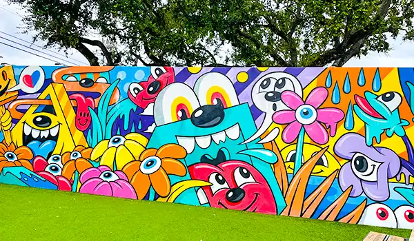 Arte de grafite colorido com desenhos de personagens divertidos, flores e elementos vibrantes em um mural ao ar livre sob árvores.