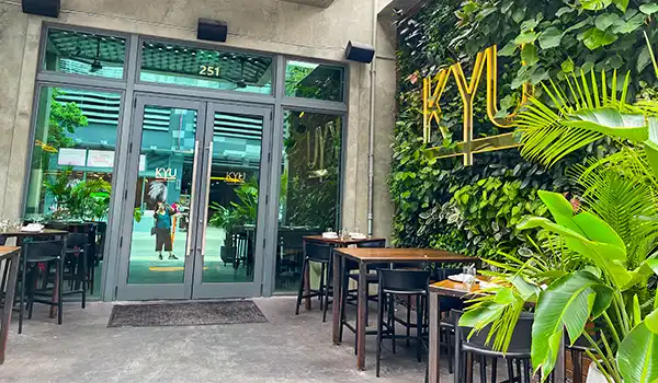 Entrada de um restaurante com cadeiras e mesas ao ar livre, decorado com jardim vertical e sinalização com a palavra KYL em destaque na parede, ideal para quem busca uma experiência gastronômica na cidade.