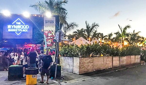 Ambiente de festa ao ar livre na Wynwood Marketplace, com pessoas se divertindo ao pôr do sol, iluminação colorida, decoração vibrante e palmeiras ao redor.