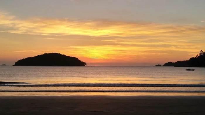 Cenário de praia ao pôr do sol com ilha ao fundo, céu dourado e mar tranquilo, ideal para momentos de relaxamento e contemplação da natureza.
