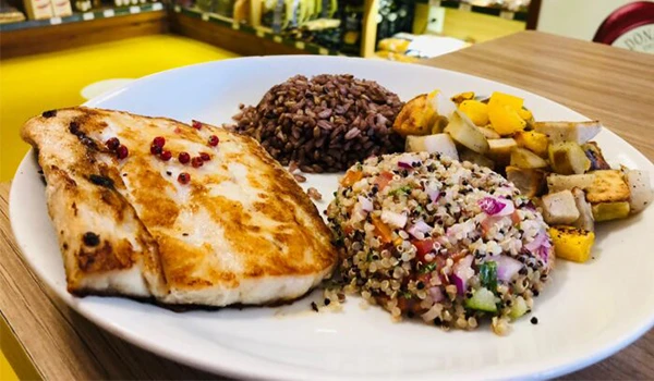 Prato de comida com filé de peixe grelhado, arroz preto, salada de quinoa e legumes assados, servido em restaurante.