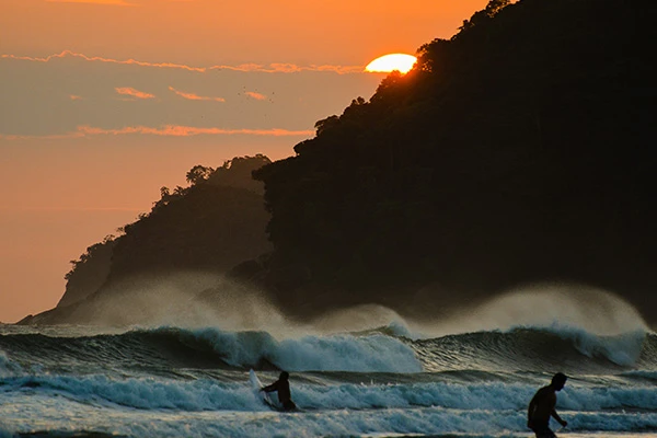 Pôr do sol na praia com surfistas aproveitando as ondas ao pôr do sol, cenário de natureza exuberante e mar tranquilo, perfeito para surfistas.