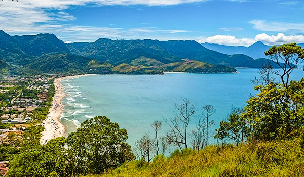 Paisagem de uma praia com água azul clara, cercada por montanhas verdes e árvores na margem, sob céu parcialmente nublado, ideal para turismo sustentável e ecoturismo.