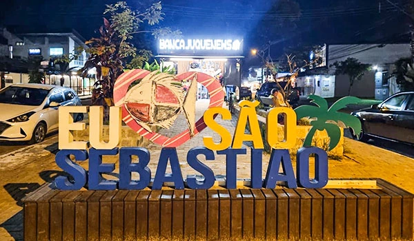 Placa decorativa com letras azuis e amarelas que dizem 'EU SÃO SEBASTIÃO', em frente à uma árvore e prédios, com iluminação noturna, simbolizando orgulho e identidade local