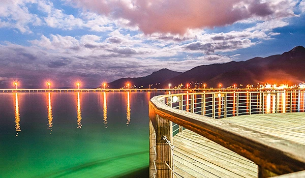 Imagem de um píer de madeira com guarda-corpo de metal ao longo de um lago tranquilo ao entardecer, com céu ensolarado e montanhas ao fundo, refletindo na água.