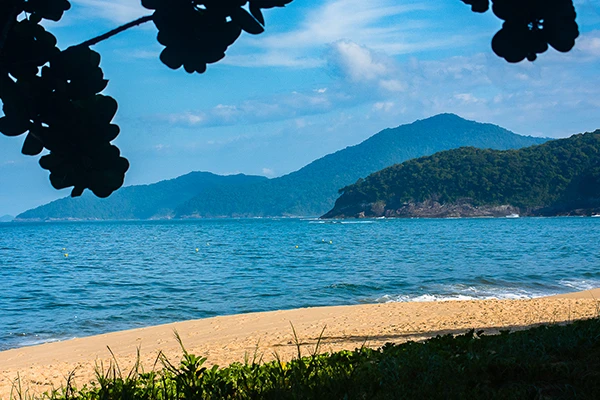 Paisagem de praia com areia, água azul e uma cordilheira ao fundo, sob céu parcialmente nublado, perfeita para descanso e lazer no litoral.