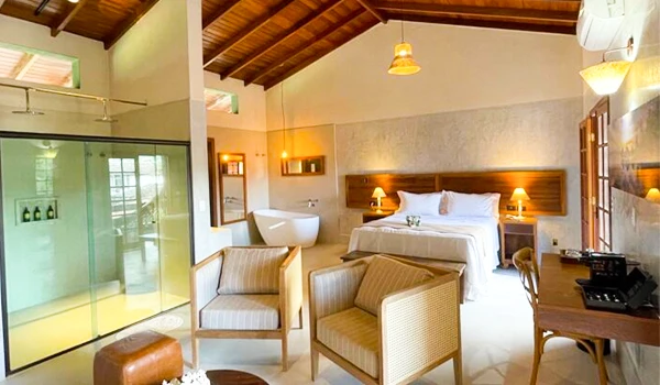 Imagem de um quarto de hotel com decoração aconchegante e iluminação suave, incluindo cama de casal, poltronas, bancada de trabalho e banheiro elegante, ideal para férias ou viagens de lazer.