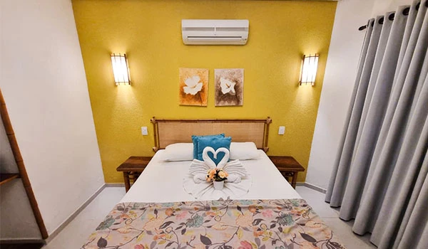 Quarto de hotel decorado com parede amarela, cama com colcha floral, vasos de flores, quadro de arte na parede, cortinas cinza ao lado, iluminação moderna e ar-condicionado