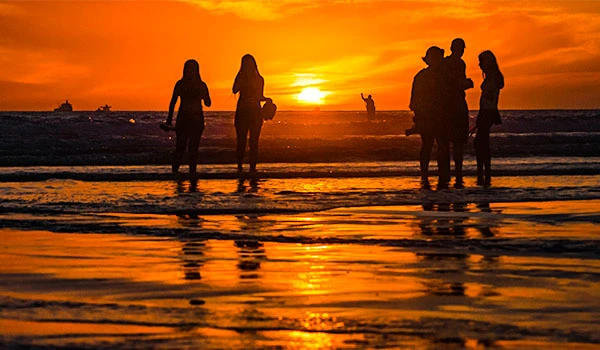 Grupo de pessoas aproveitando o pôr do sol na praia ao entardecer, com o mar refletindo as cores quentes do céu laranja e amarelo, criando uma atmosfera tranquila e relaxante.