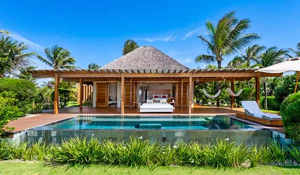Vista frontal de um bangalô de luxo com piscina privativa em primeiro plano. A construção tem estrutura de madeira e telhado de palha, cercada por um deck com espreguiçadeiras, redes e jardins tropicais com coqueiros. O céu está limpo e azul, transmitindo uma atmosfera de tranquilidade e exclusividade