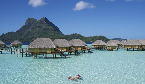 Bangalôs de palha sobre águas cristalinas em Bora Bora, com uma pessoa nadando no primeiro plano e uma montanha verde ao fundo sob céu azul.