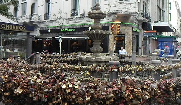 Vizinhança de rua em Montevideu com uma fonte e várias cadeados presos na grade, simbolizando amor e compromisso, em uma área movimentada com prédios históricos ao fundo.
