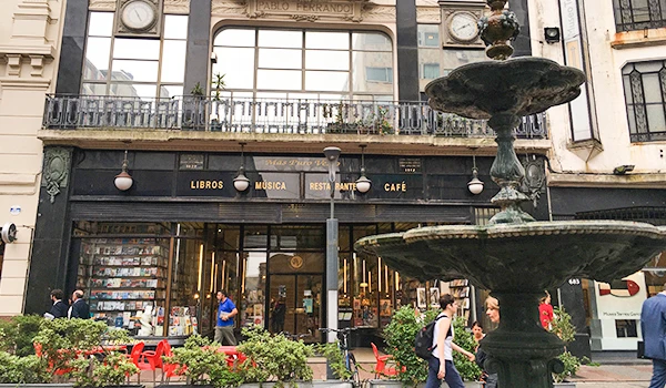 Imagem de um prédio com fachada de vidro, contendo uma livraria, restaurante, bar e cafeteria, com pessoas caminhando na rua e uma fonte de ferro na frente, cenário urbano na cidade  de Montevideu .
