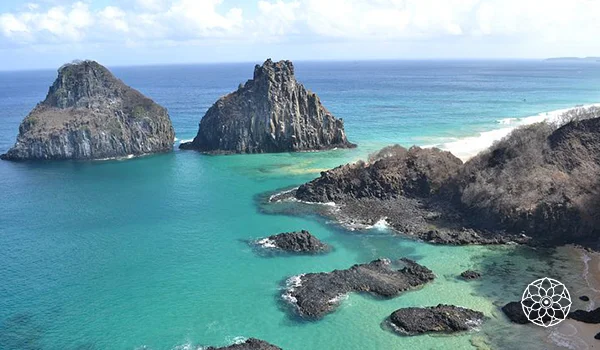 Vista aérea de uma costa tropical com águas azul-turquesa, formações rochosas e duas ilhotas escarpadas próximas à praia de areia clara.