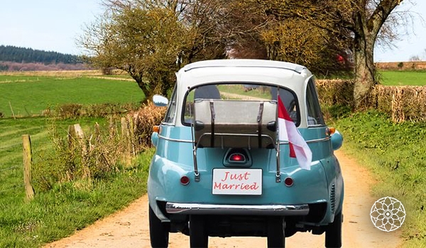 Carro antigo azul-claro com a inscrição “Just Married”, decorado com bandeiras, percorrendo uma estrada rural cercada por campos e árvores.