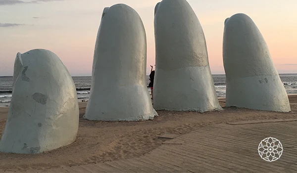 Imagem de esculturas na praia que parecem dedos gigantes de uma mão, com pôr do sol ao fundo, criando uma cena artística e única.