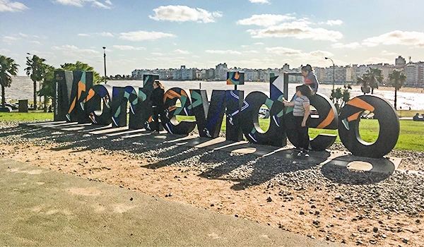 Pessoas em atividade ao redor do letreiro Montevideu, na orla da cidade com vista para o rio, durante o dia. A cena captura o movimento e a energia urbana, uma das atrações de Montevidéu.