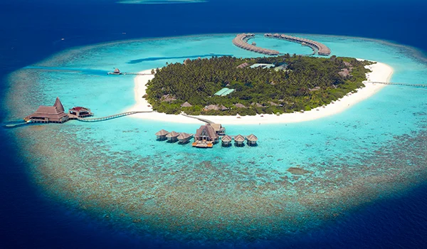 Vista aérea de uma ilha tropical com areia branca, vegetação densa e águas azul-turquesa, cercada por bangalôs sobre o mar e um píer curvo ao fundo.