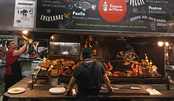 Cozinha  do Mercado em Montevideu, com variedade de carnes, frangos e porcos assados, focada em carne, frango, porco e aves, com ambiente de restaurante ao fundo.