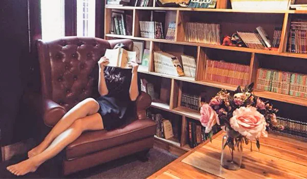 Mulher relaxando em uma poltrona enquanto lê um livro em um ambiente aconchegante, com estante de livros e flores ao fundo. Dicas para presente quem gosta de viajar.
