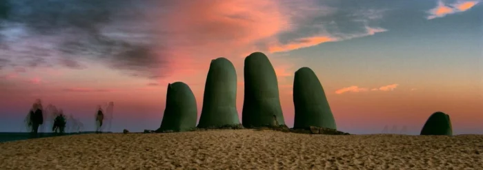 Imagem da inscrição Olho no Olho em frente ao pôr do sol, em um local com areia, com algumas pessoas ao fundo, na cidade de Brasília, com destaque para as esculturas de mãos na obra de arte moderna.