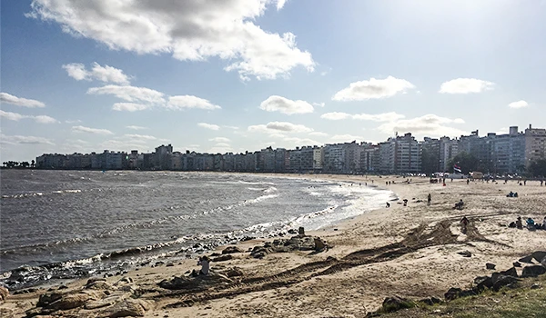 Rambla à Beira do Rio da Prata, com vista para o mar e prédios no horizonte, céu ensolarado com algumas nuvens, ambiente de lazer e descanso à beira-mar. Um dos destaques sobre o que fazer em Montevideu.