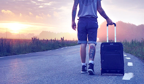 Homem caminhando em uma estrada com uma mala, ao fundo um lindo pôr do sol, representando a preparação para viagens e aventuras. Dicas para presente quem gosta de viajar.