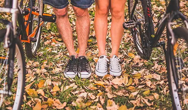 Casal de pé sobre o chão coberto de folhas secas, usando tênis casual, com bicicletas ao lado, ideal para passeios ao ar livre. Dicas para presente quem gosta de viajar.