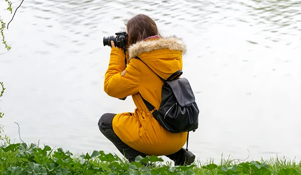 Mulher vestindo um casaco amarelo fotografando com uma câmera à beira de um lago, cercada por grama verde. Dicas para presente quem gosta de viajar.