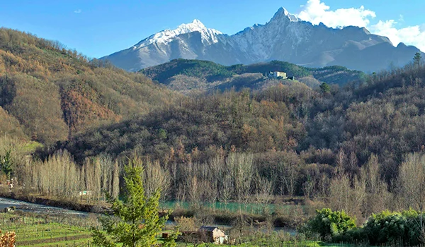 Paisagem rural com vegetação e colinas em primeiro plano e uma cadeia de montanhas escarpadas sob céu azul ao fundo na Lua de Mel na Toscana.